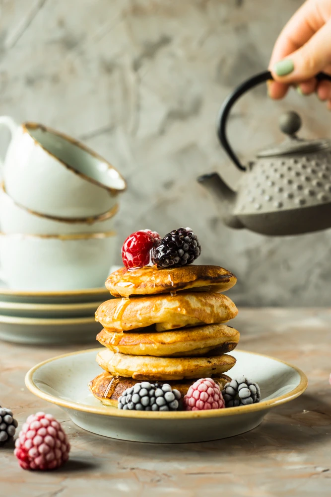 A stack of golden homemade pancakes topped with honey and fresh berries, with a teapot pouring in the background.