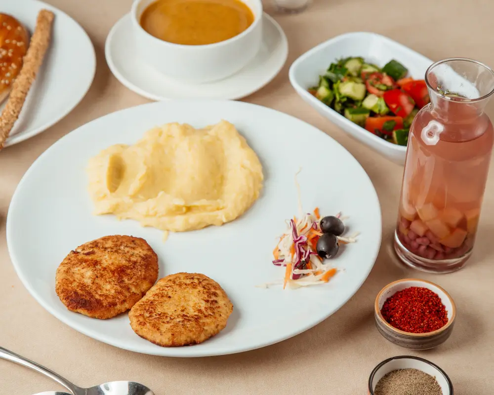 A plate of mashed potatoes and two crispy patties served with a small salad, olives, and coleslaw, accompanied by soup, breadsticks, and a glass of fruit-infused water