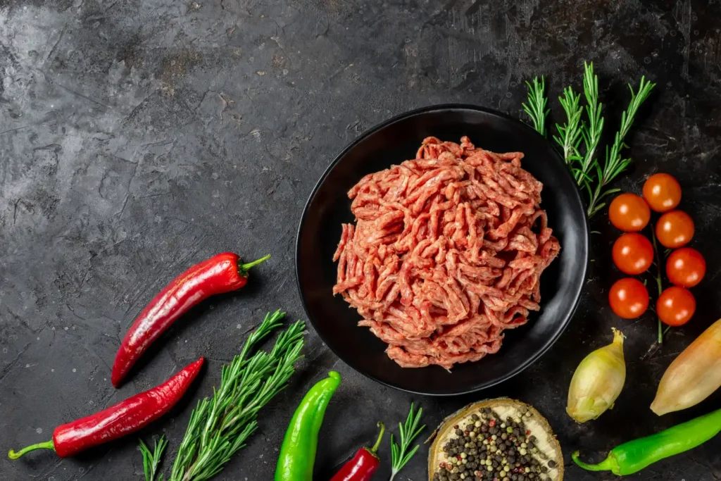 Ground chicken in a black bowl surrounded by fresh rosemary, chili peppers, cherry tomatoes, onions, and peppercorns on a dark background.