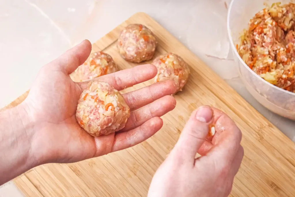 Hands shaping chicken ricotta meatballs on a wooden cutting board with a bowl of the meat mixture in the background.