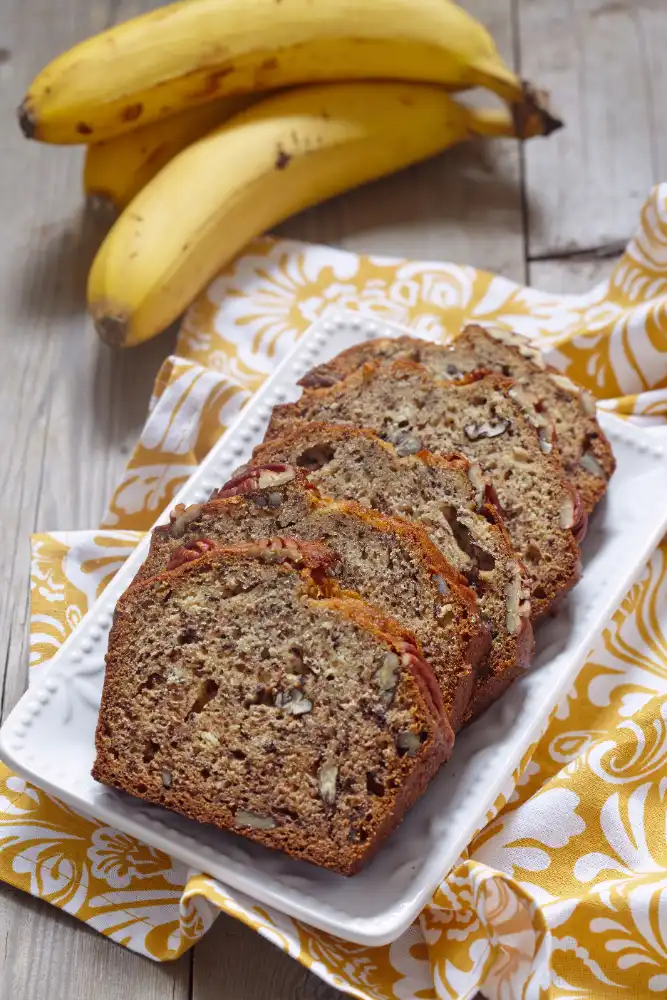 Sliced banana bread with nuts on a white plate, with fresh bananas in the background.