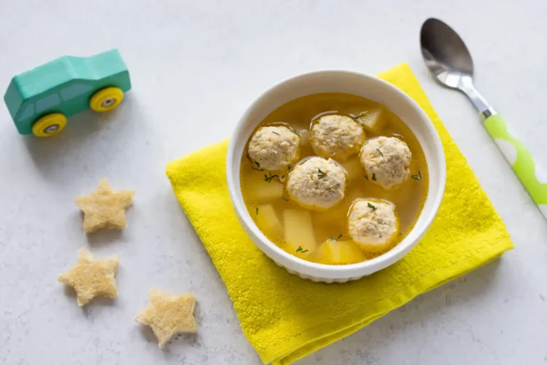 A bowl of soup with meatballs and diced potatoes on a yellow napkin, accompanied by a toy car, star-shaped bread, and a child’s spoon.