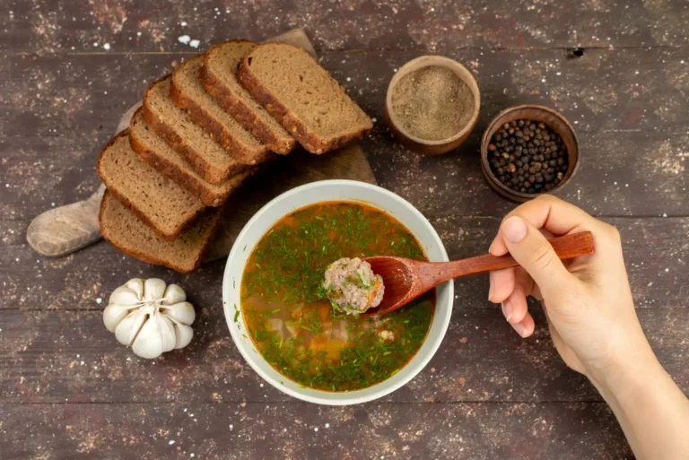 A bowl of meatball soup garnished with fresh herbs, accompanied by slices of rye bread, garlic, and spices on a rustic wooden table