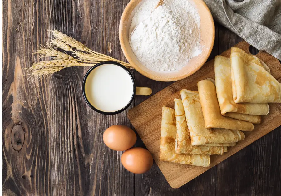 Flat lay of pancake ingredients, including flour, milk, eggs, and freshly prepared crepes, on a rustic wooden table.