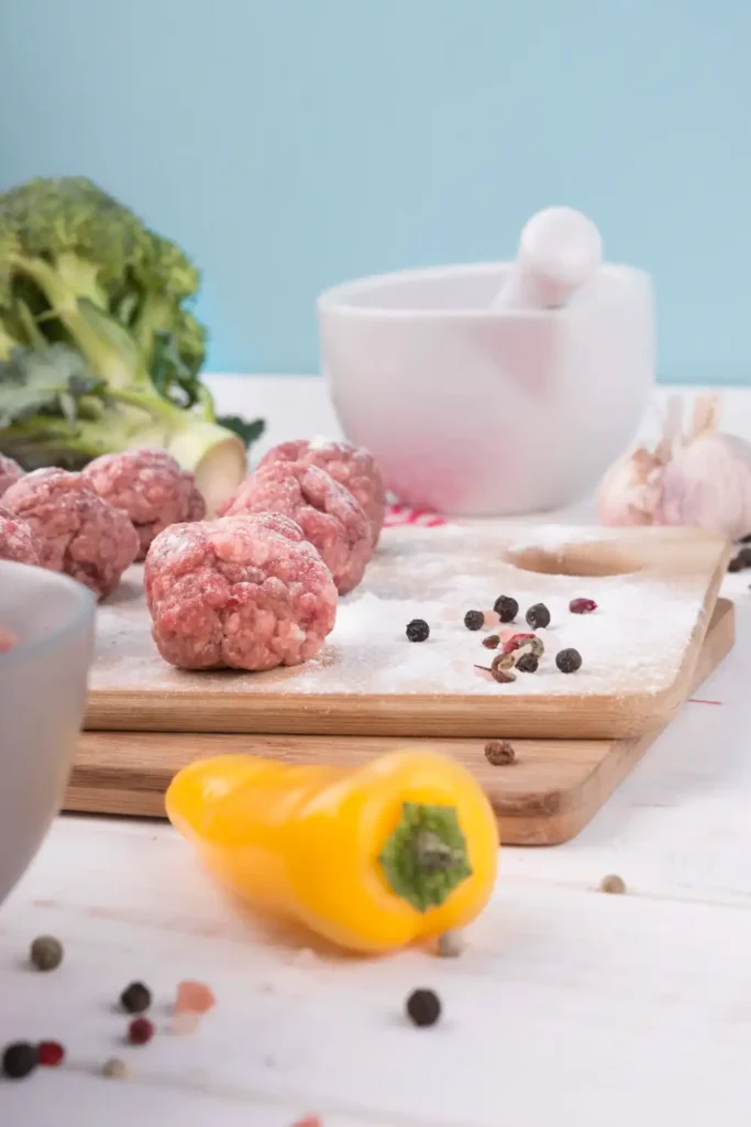 Uncooked chicken ricotta meatballs on a floured cutting board with fresh vegetables and spices in the background