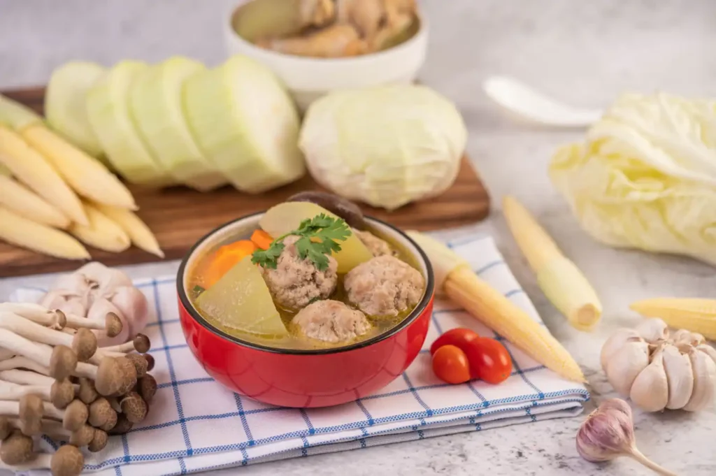 A vibrant bowl of meatball soup with sliced winter melon, mushrooms, baby corn, and fresh vegetables on a checkered cloth, surrounded by ingredients like garlic, cabbage, and tomatoes.