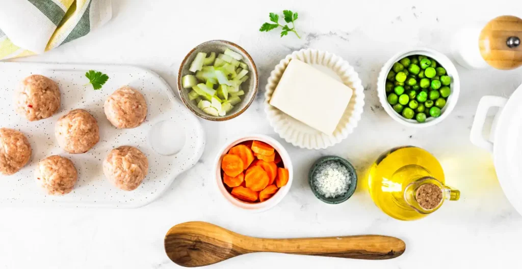 A flat lay of raw chicken meatballs and ingredients including carrots, celery, peas, butter, and olive oil on a white countertop.