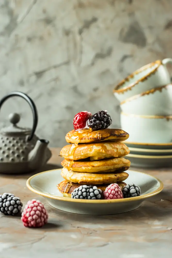 A stack of fluffy homemade pancakes topped with blackberries and raspberries, drizzled with honey, with a rustic teapot and ceramic dishes in the background.