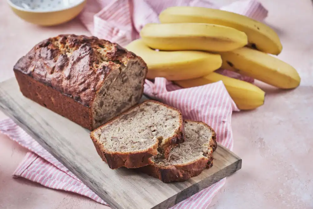 Whole banana bread loaf with slices on a wooden board, surrounded by fresh bananas.