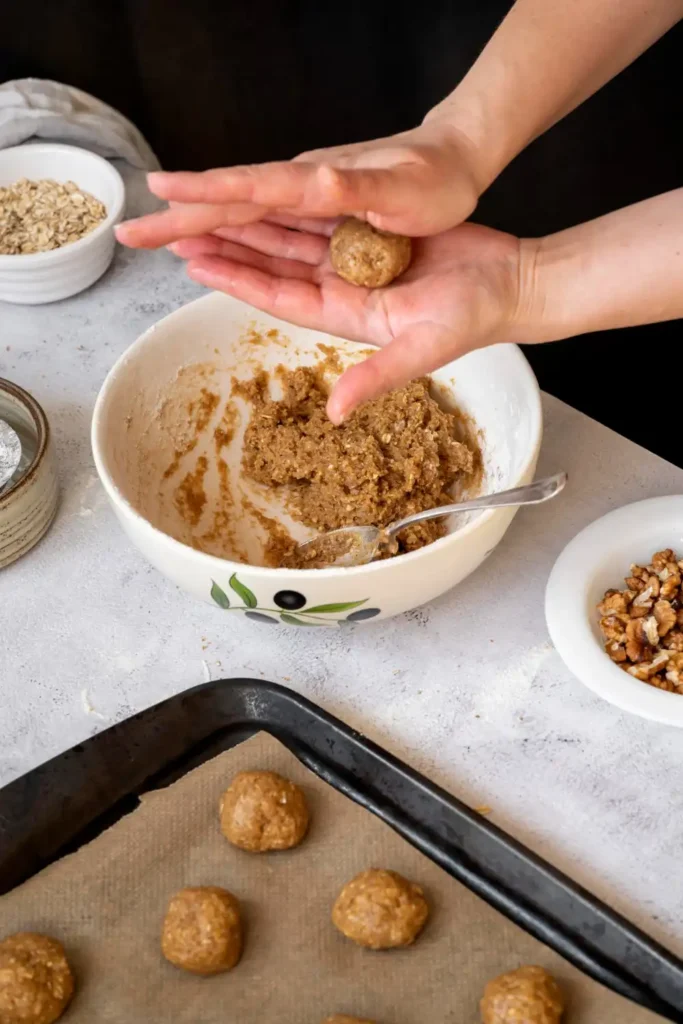 Hands rolling oat and nut mixture into small balls on a baking tray lined with parchment paper, surrounded by bowls of ingredients like walnuts and oats.