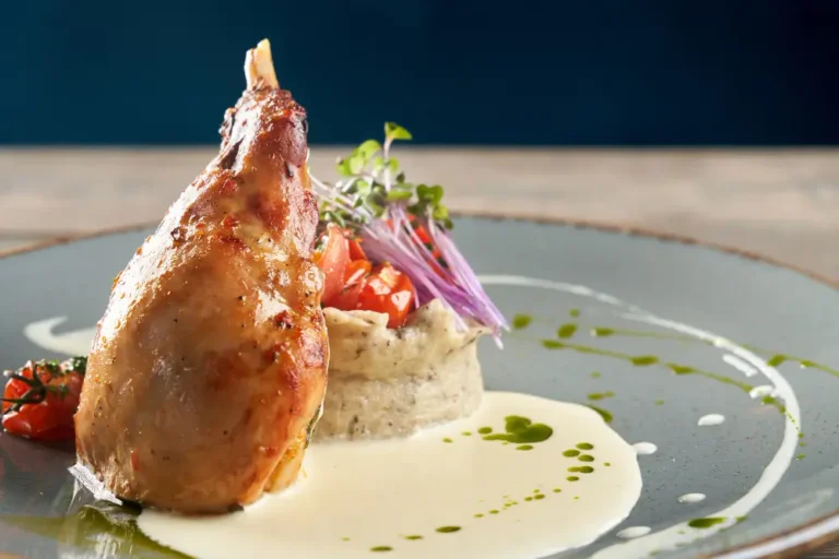 A beautifully roasted chicken leg served with a creamy sauce, mashed potato garnish, sautéed tomatoes, microgreens, and edible flowers on a sophisticated plate.