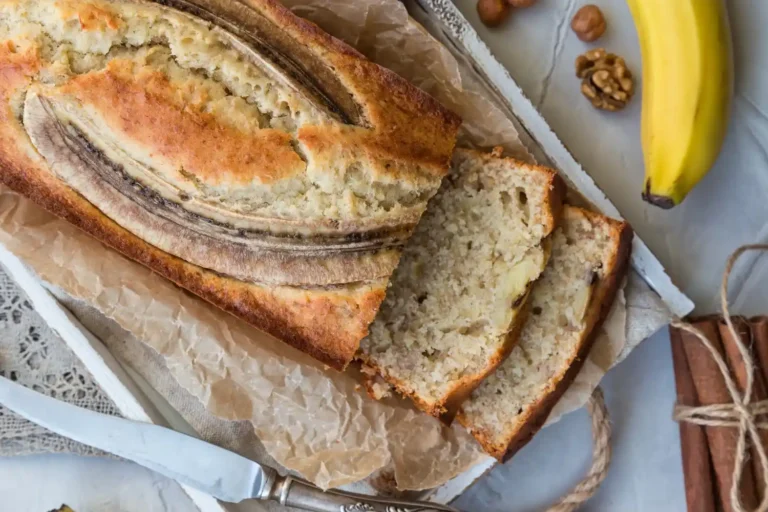 A golden-brown loaf of banana bread with a sliced top, surrounded by a fresh banana, walnuts, and cinnamon sticks on a rustic tray.