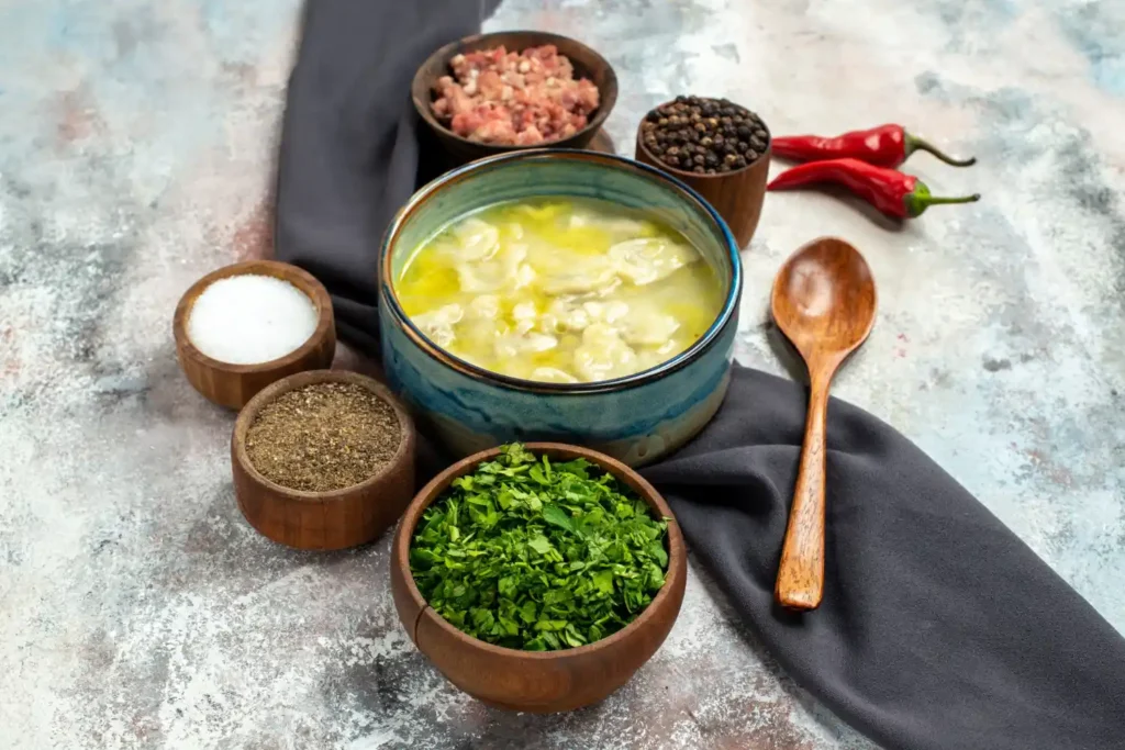 A bowl of chicken broth surrounded by fresh herbs, spices, and seasonings on a rustic table.