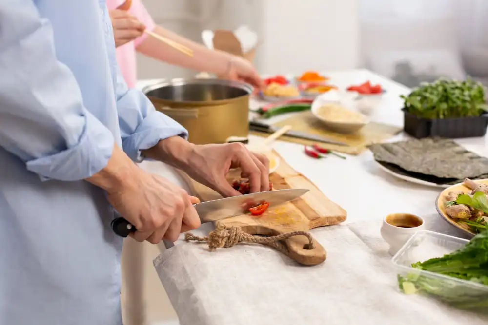 A person chopping fresh tomatoes on a wooden cutting board, surrounded by other cooking ingredients, including vegetables, herbs, and sauces, on a white kitchen table.