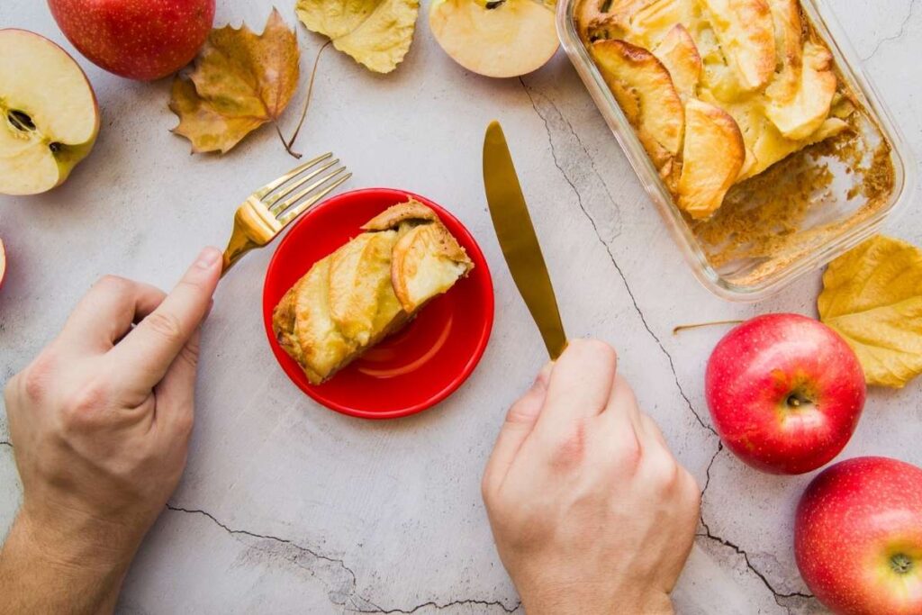 A slice of homemade apple cake served on a red plate with fresh apples and autumn leaves.