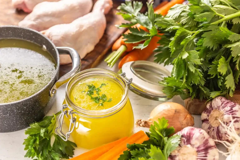 A pot of chicken broth alongside fresh vegetables, herbs, and raw chicken pieces.