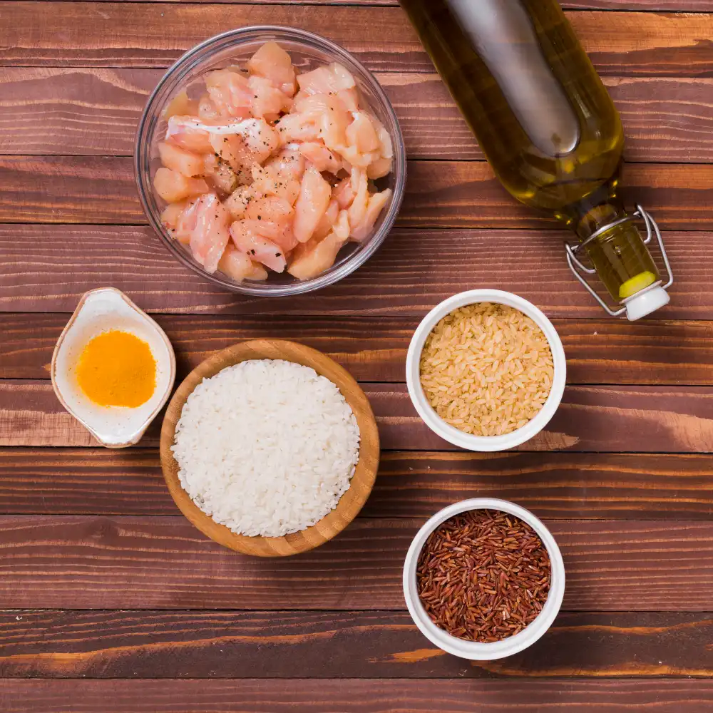 An overhead shot of chicken pieces in a bowl, olive oil, rice, red rice, brown rice, turmeric powder, and seasoning arranged on a wooden table.