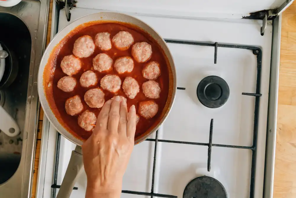 A hand carefully arranging raw chicken meatballs in a skillet filled with tomato sauce, on a stovetop.