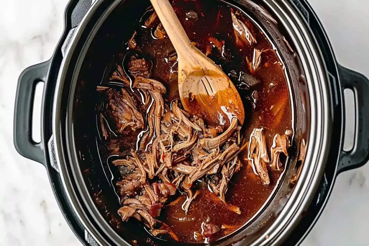 A slow cooker filled with tender, shredded beef simmering in a rich, flavorful birria broth, stirred with a wooden spoon.