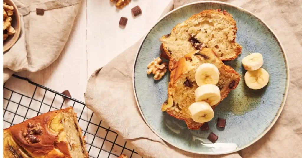 Two slices of banana bread served on a plate with fresh banana slices and walnuts.