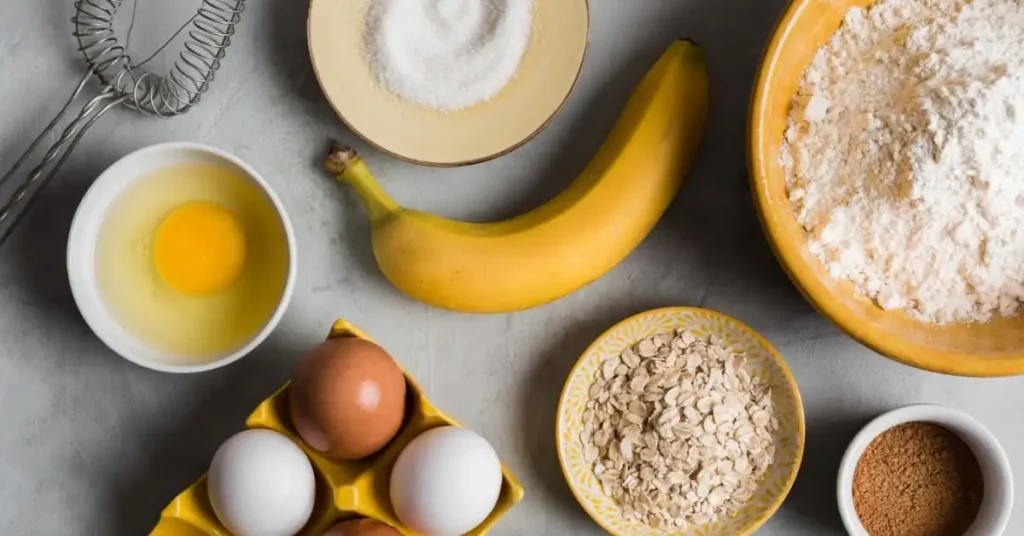 Flat lay of banana bread ingredients including a ripe banana, eggs, flour, sugar, and oats on a gray countertop.