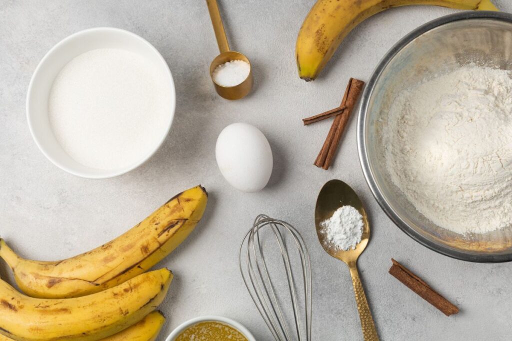 Banana bread ingredients including ripe bananas, flour, sugar, an egg, baking powder, cinnamon sticks, and salt, arranged on a gray background.