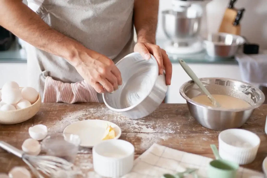 A baker greasing a baking pan with butter, surrounded by bowls of batter, eggs, and baking utensils on a wooden countertop.