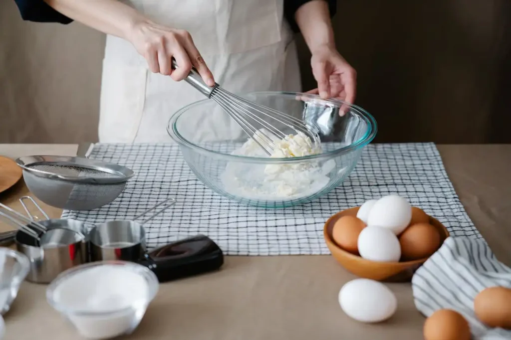 Hands whisking batter in a glass bowl with baking ingredients on the table.